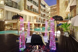 A rooftop pool area with a table set for two, surrounded by decorative lights and a flower arrangement.