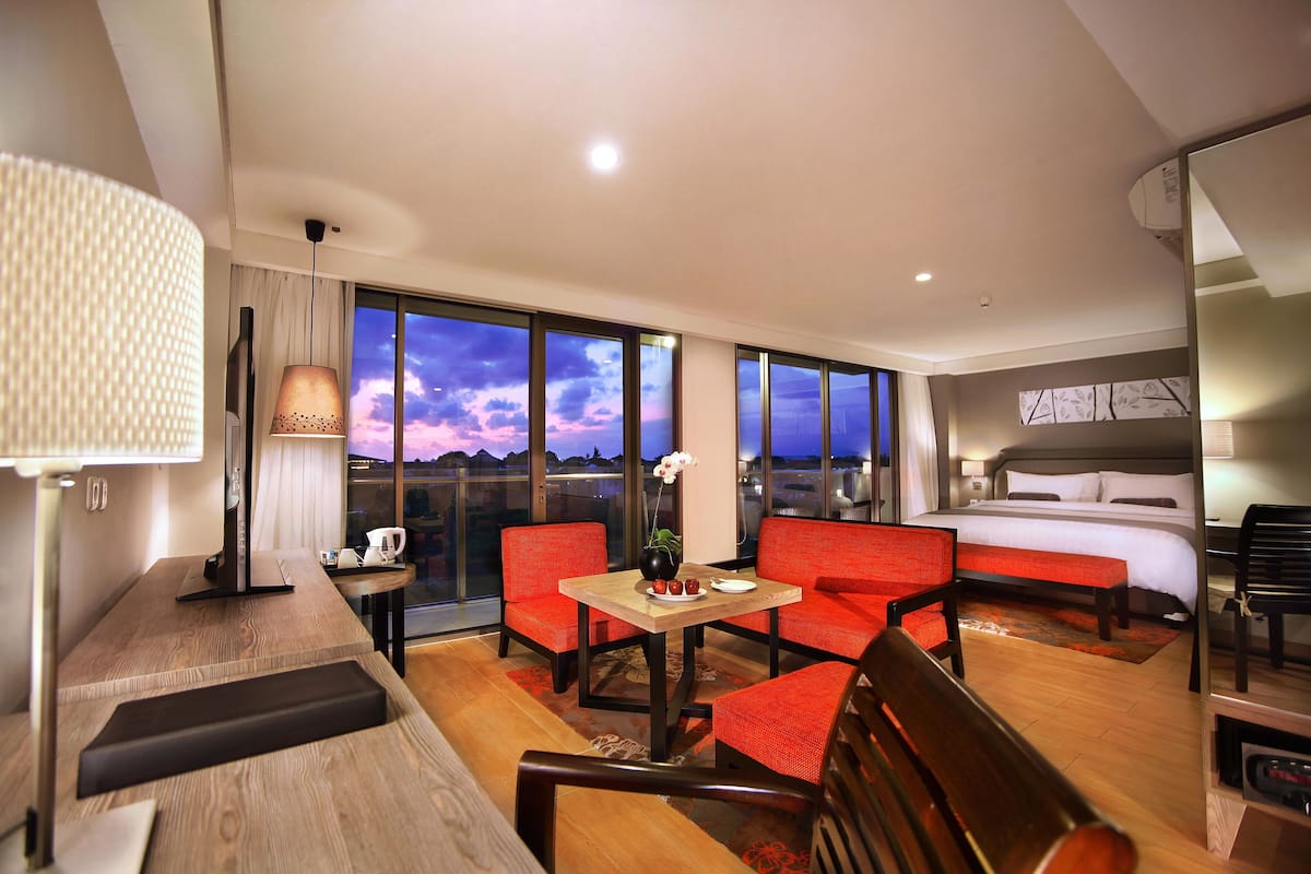 A hotel room with a bed, a desk with a lamp, two red chairs, a small table with a vase, and a large window with a city view.