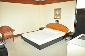 Une chambre d’hôtel avec un lit simple, une tête de lit en bois, une chaise en osier, une table de chevet avec une lampe et un petit bureau avec un téléphone.