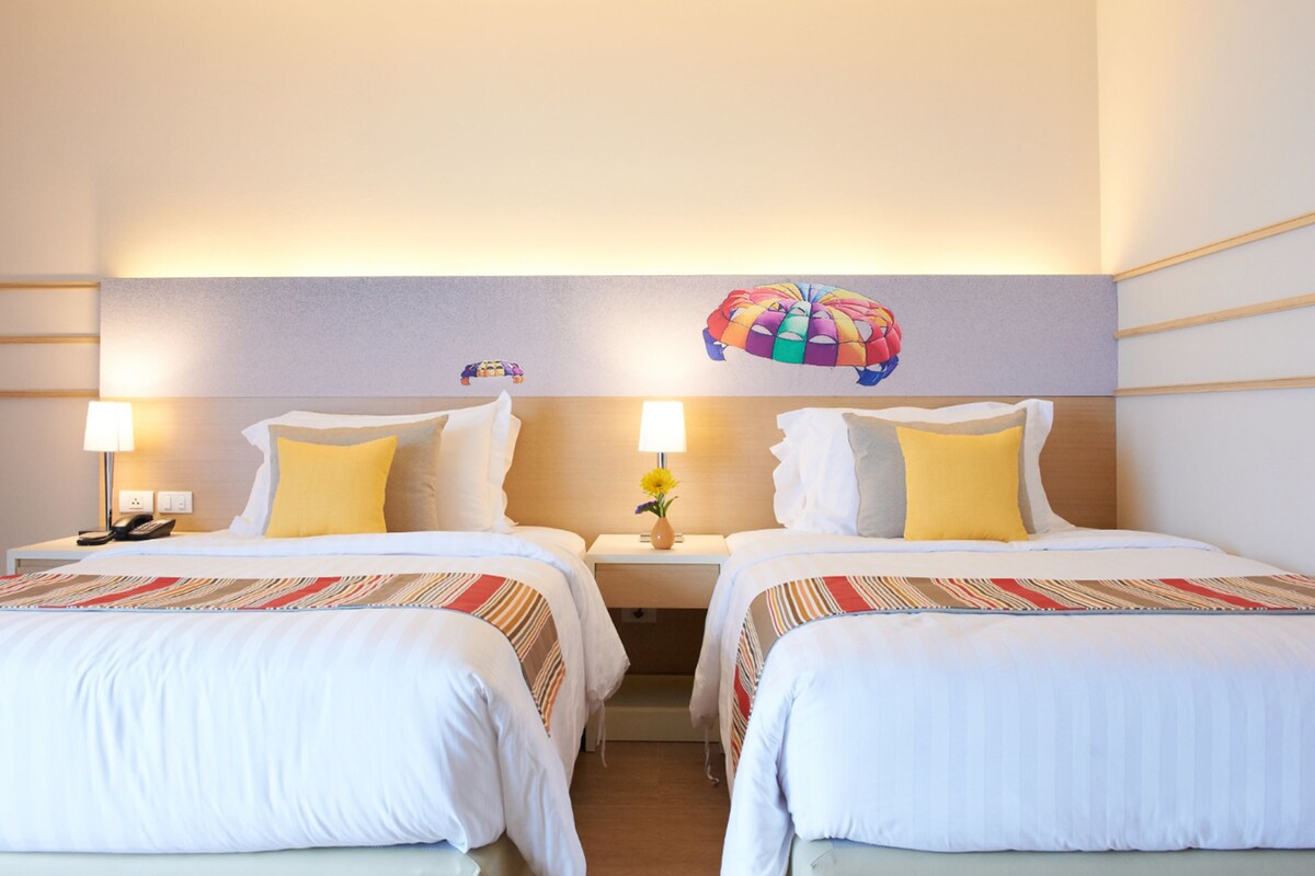 A hotel room with two beds, each with a striped bedspread, a nightstand with a lamp, and a wall decoration of a colorful parachute.