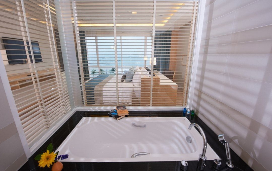 A bathroom with a large bathtub, a window with blinds, and a view of a pool and deck area.
