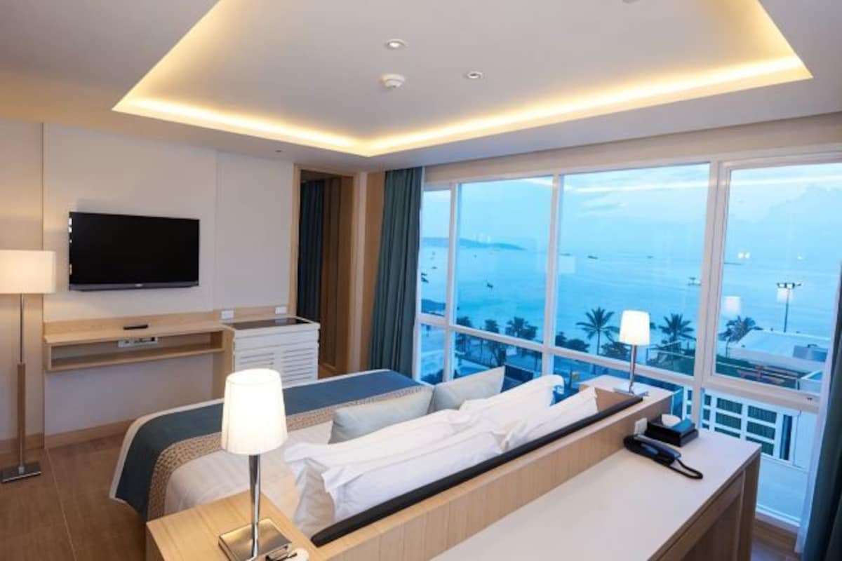A hotel room with a bed, desk, and a large window offering a view of the sea.