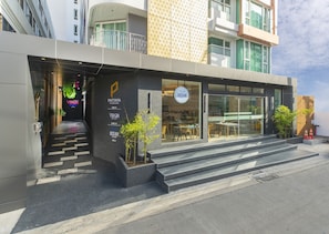 Hotel moderno com fachada de vidro, área de estar externa e escadaria que leva à entrada.