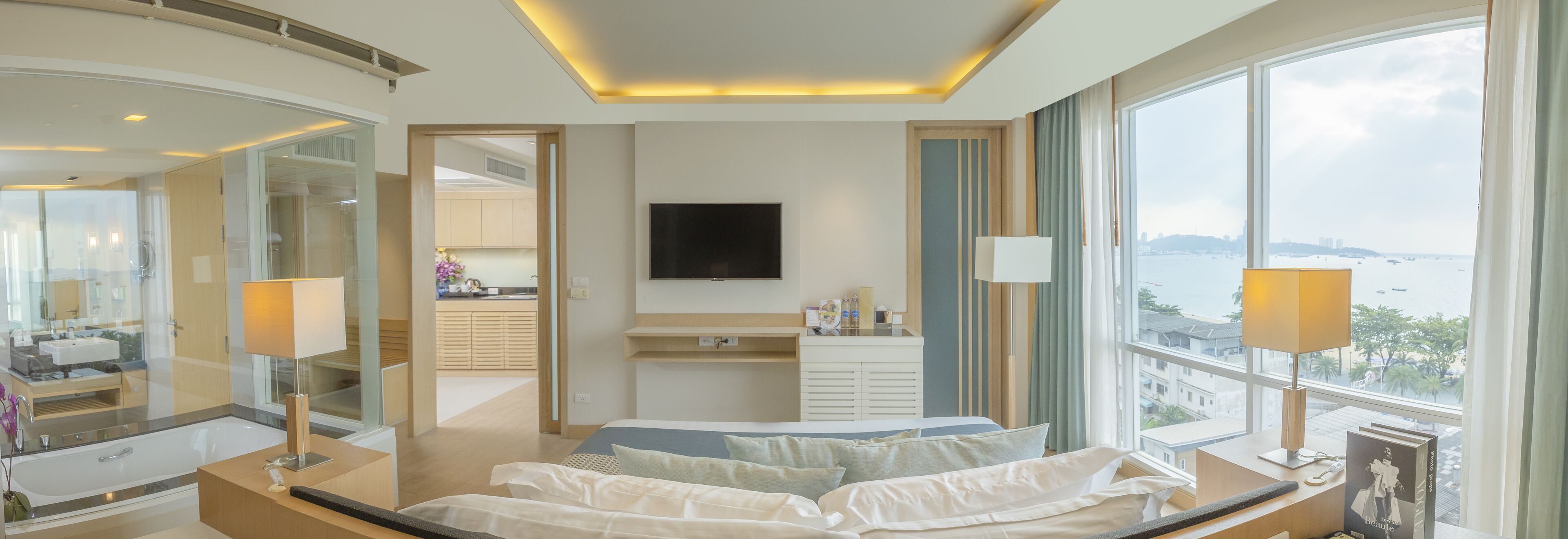 A modern hotel room with a large bed, a TV mounted on the wall, a view of the sea, and a bathroom with a bathtub and vanity.