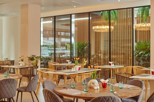 Restaurante com mesas redondas, cadeiras de madeira e vista para o verde através de grandes janelas.