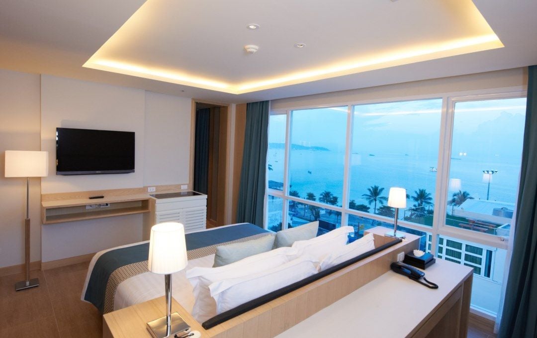 A hotel room with a bed, bedside table, lamp, TV, and a large window with a view of the sea.