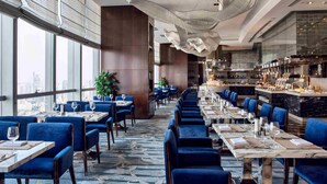 Un restaurant moderne avec de longues tables, des chaises à l’assise rembourrée de tissu bleu et un grand luminaire au plafond au design abstrait.