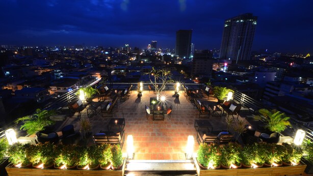 A rooftop terrace with outdoor seating, potted plants, and city lights in the background.