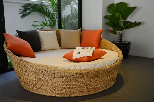 A round woven wicker sofa with a cushion, placed in a room with a large potted plant and a window with a view of greenery. - Kolab Sor Phnom Penh Hotel (Phnom Penh)