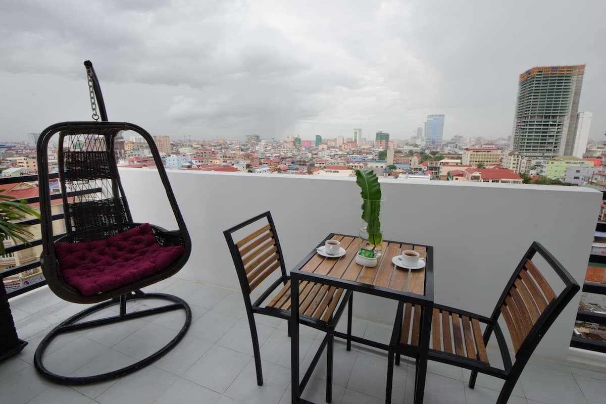 Un balcón con una mesa y sillas de madera, una silla acolchada y vistas a un paisaje urbano con edificios.