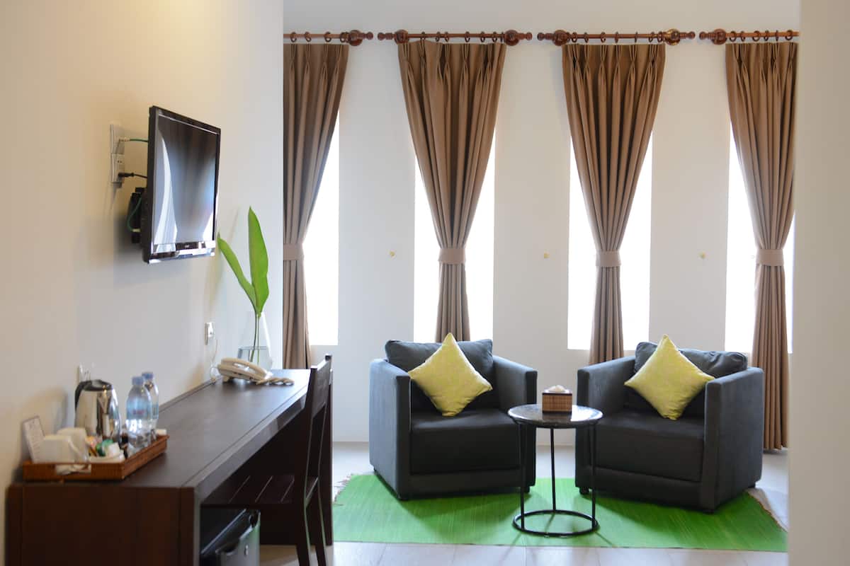 Habitación de hotel con dos sillones oscuros, una mesita redonda pequeña, un escritorio con teléfono y una bandeja de objetos, y un ventanal con cortinas.