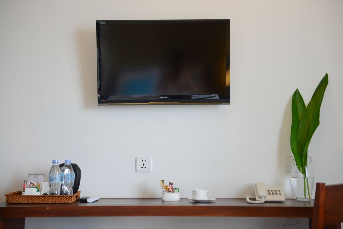 Un televisor de pantalla plana montado en una pared blanca, un escritorio de madera con un cesto de artículos de aseo, un jarrón de cristal con una planta y un teléfono.
