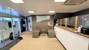A reception area with a reception desk, two wicker chairs, a small round table, and a carpeted floor. - Star Capsule Okinawa (Naha)
