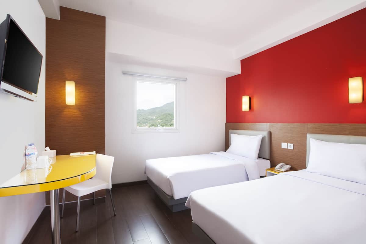 Une chambre d’hôtel avec deux lits, un bureau avec une chaise, une télévision et une fenêtre donnant sur un espace verdoyant.