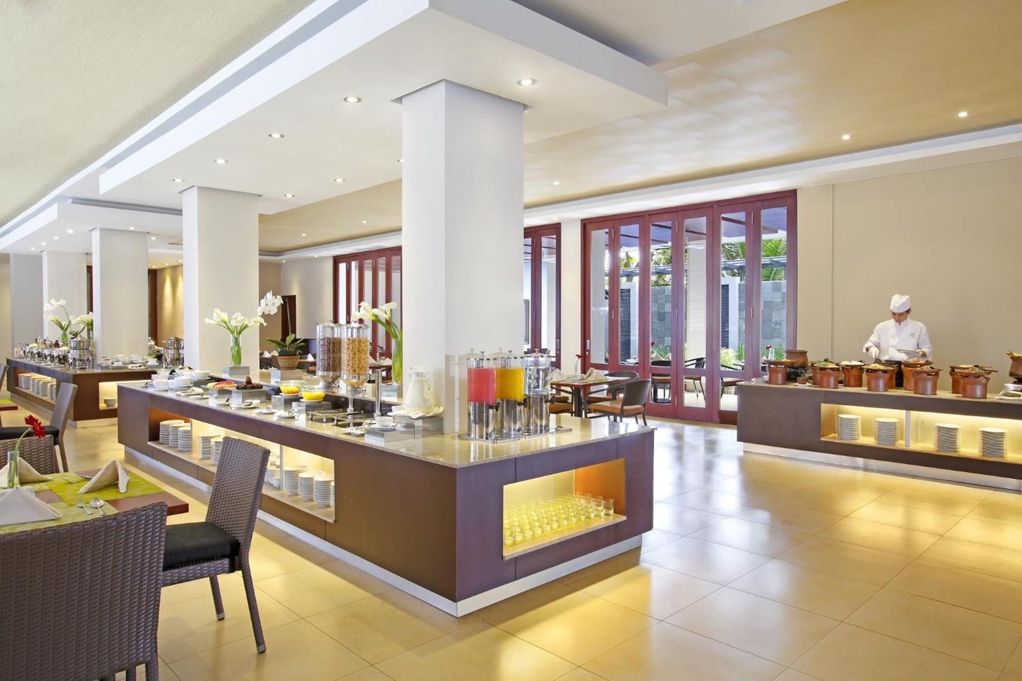 Um hall de hotel espaçoso com área de buffet central, disposição de assentos e vista para o exterior por grandes janelas.