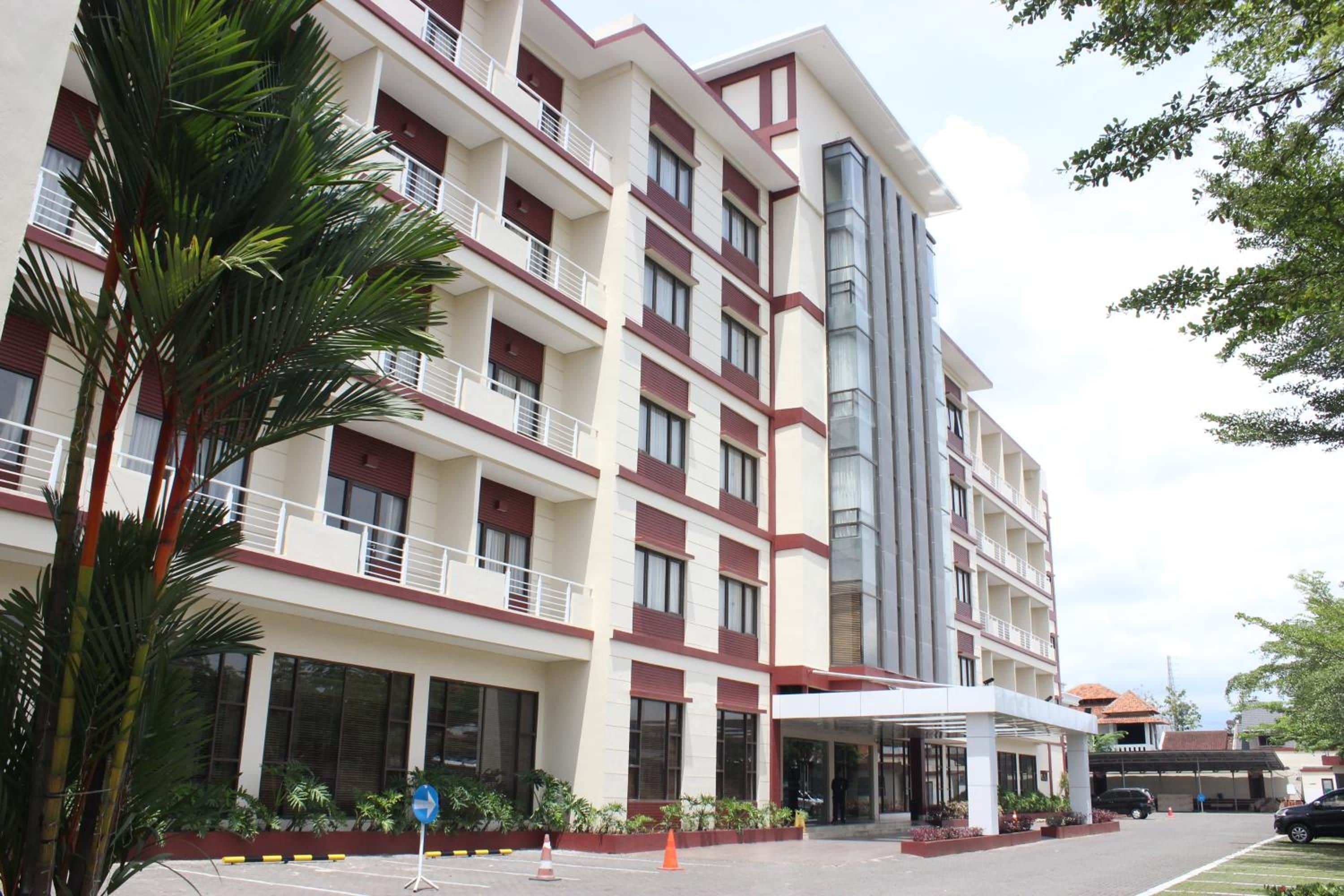 A multi-story hotel with a central entrance, balconies on each floor, and a parking area in front.