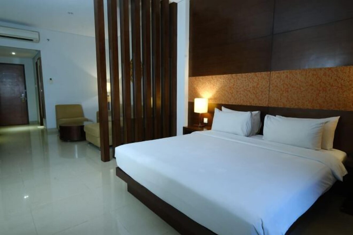 A hotel room with a large bed, a wooden partition, a chair, and a nightstand with a lamp.