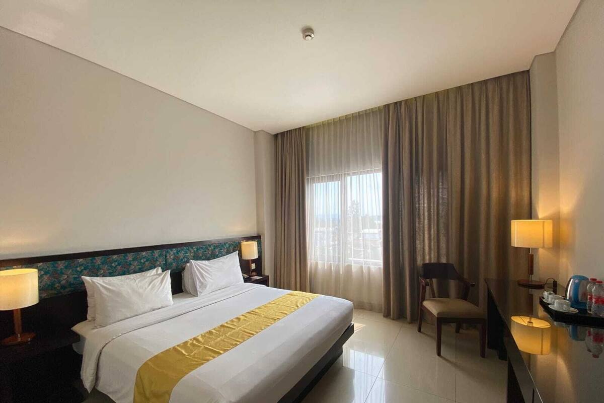 Une chambre d’hôtel avec un grand lit, un bureau avec une chaise, une lampe et une vue sur l’extérieur par une fenêtre munie de rideaux.