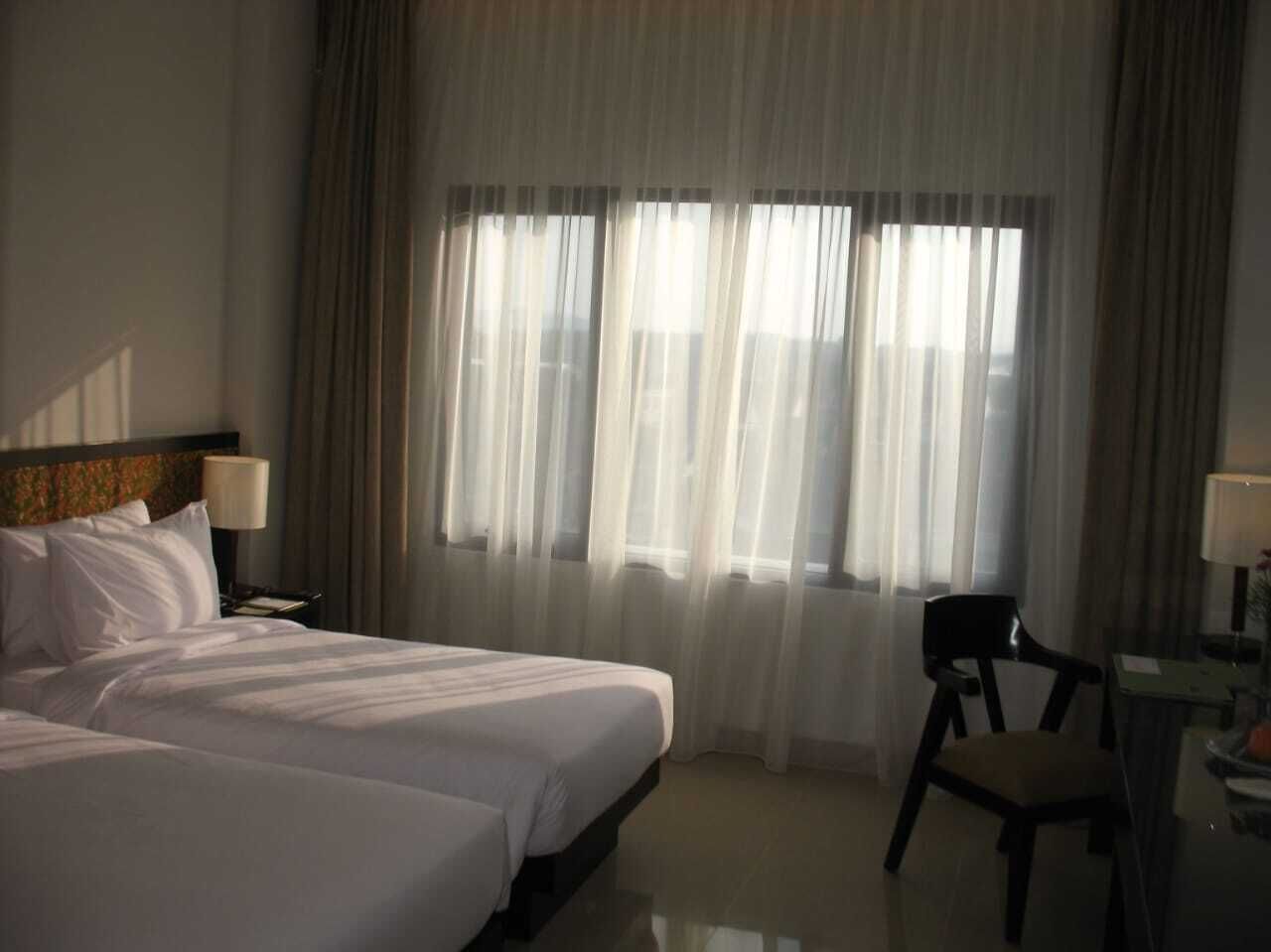 A hotel room with two beds, a chair, a desk, and a window with sheer curtains.