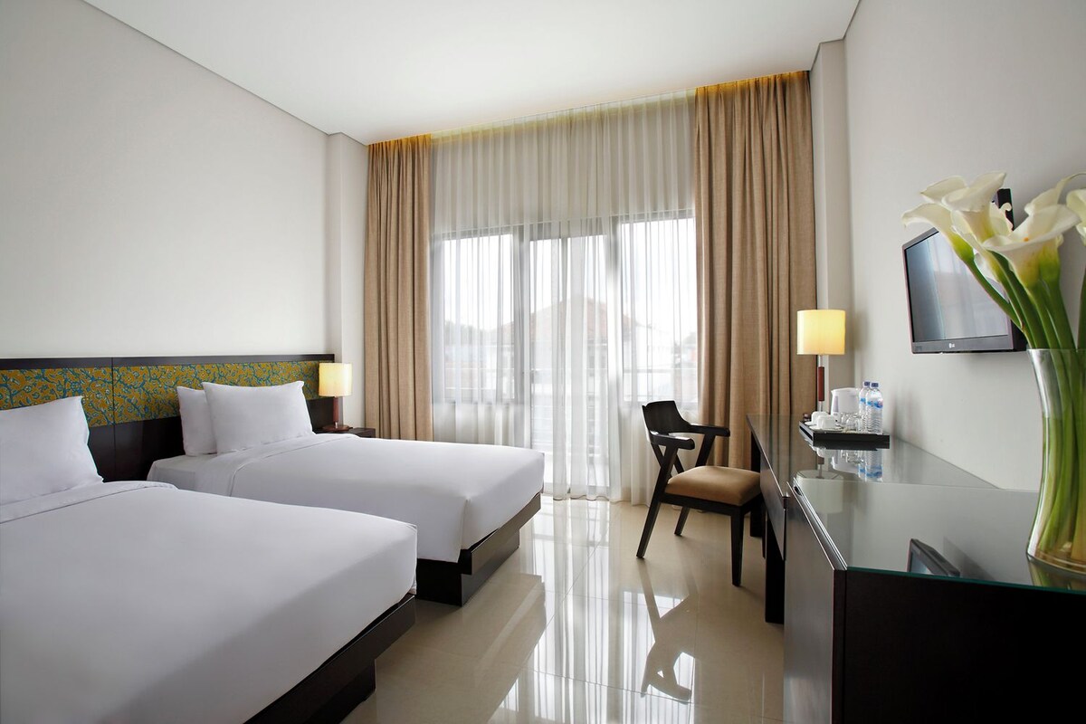 A hotel room with two beds, a desk, a chair, a TV, and a vase with flowers.