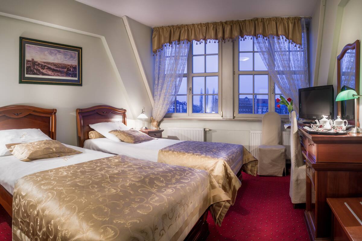 Ein Hotelzimmer mit zwei Betten, einem Schreibtisch mit Fernseher und einem Fenster mit Vorhängen.