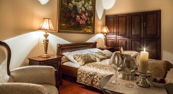 Habitación de hotel con una cama, dos lámparas de noche, una mesita de noche, un cuadro floral, un armario de madera y una mesa con una vela, vasos y un frutero.