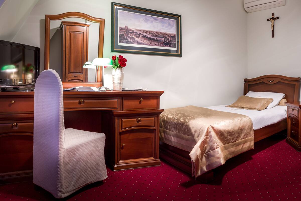 Ein Hotelzimmer mit Holztisch, Stuhl, einem Bett mit beiger Bettdecke, einem Nachttisch, einem gerahmten Bild an der Wand und einem Kruzifix an der Wand.