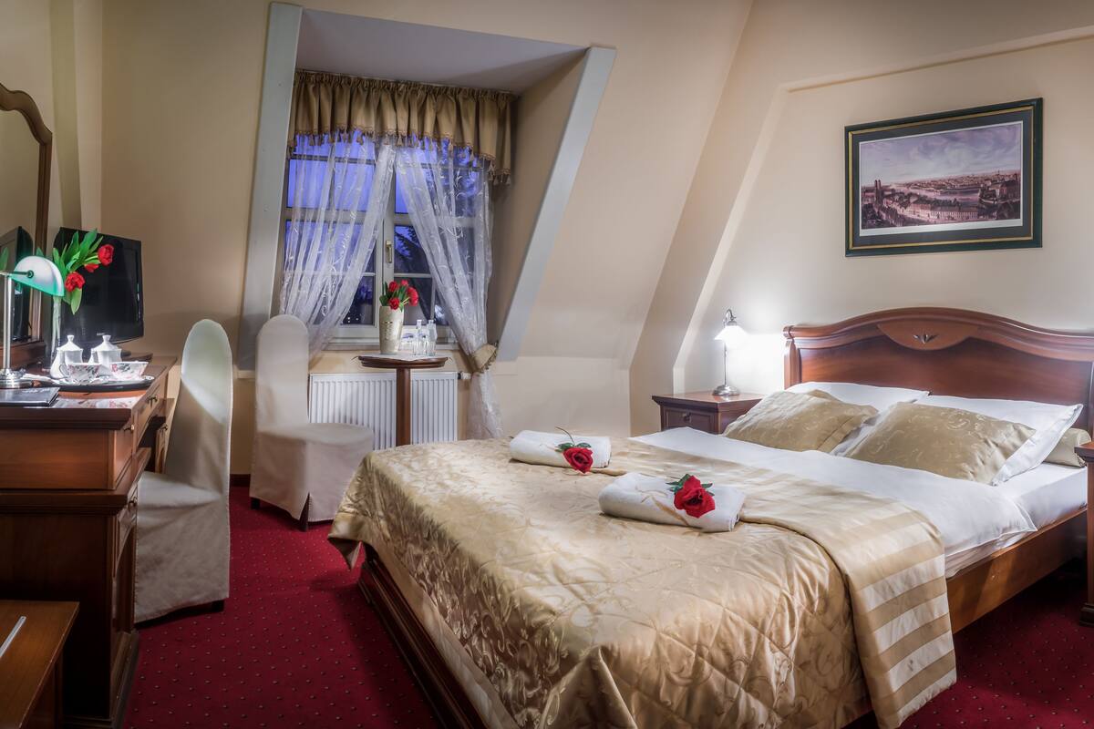 Ein Hotelzimmer mit einem großen Bett, einem Schreibtisch mit Lampe, einem Sessel, einem Fenster mit Vorhängen und einem gerahmten Bild an der Wand.