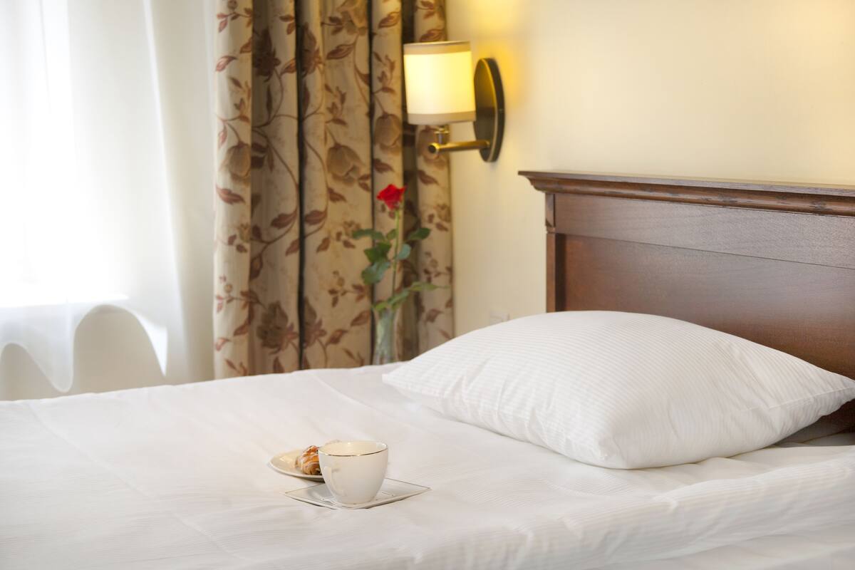 Une chambre d’hôtel avec un lit, une tasse de café sur une soucoupe, un rideau à motifs floraux et une lampe fixée au mur.