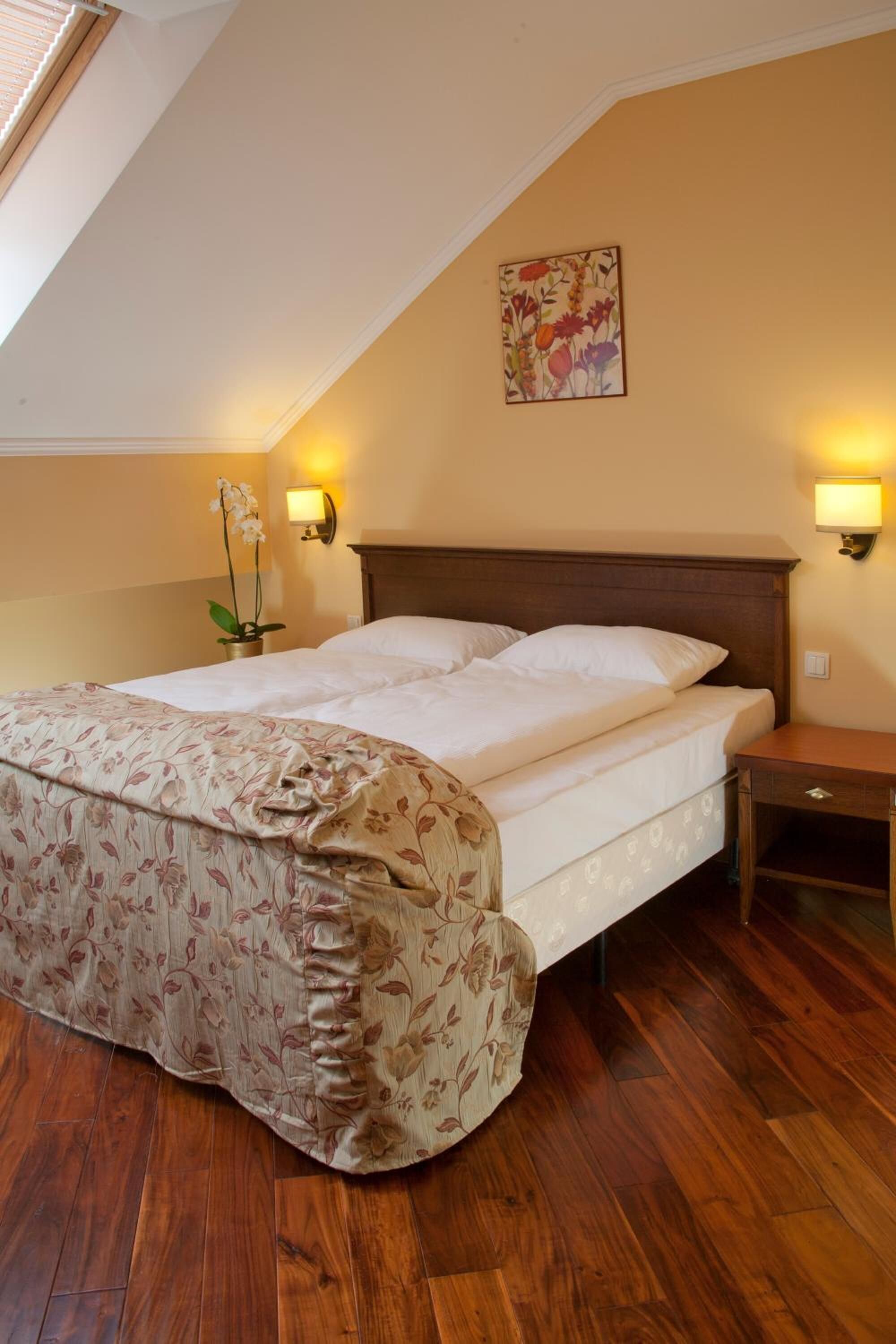 Un dormitorio con piso de madera, una cama con colcha floral, dos lámparas de pared, una mesita de noche y un cuadro en la pared.