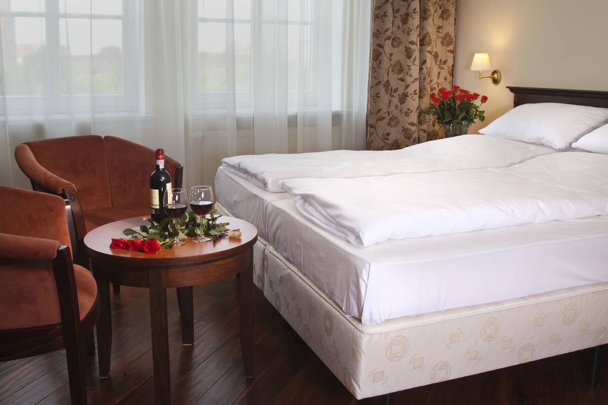 Une chambre d’hôtel avec un lit, deux chaises, une petite table avec une bouteille et des verres, et un vase avec des fleurs.