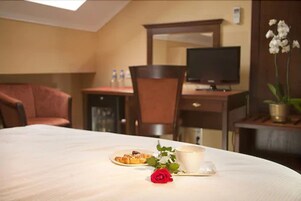 A hotel room with a dining table set with a plate of food, a cup, and a rose. There is a TV on a stand and a chair in the background.