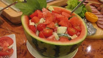 A watermelon bowl filled with a salad, surrounded by various food items on a wooden table.