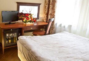 A hotel room with a bed, a desk with a computer monitor, a chair, a small refrigerator, and a window with curtains.