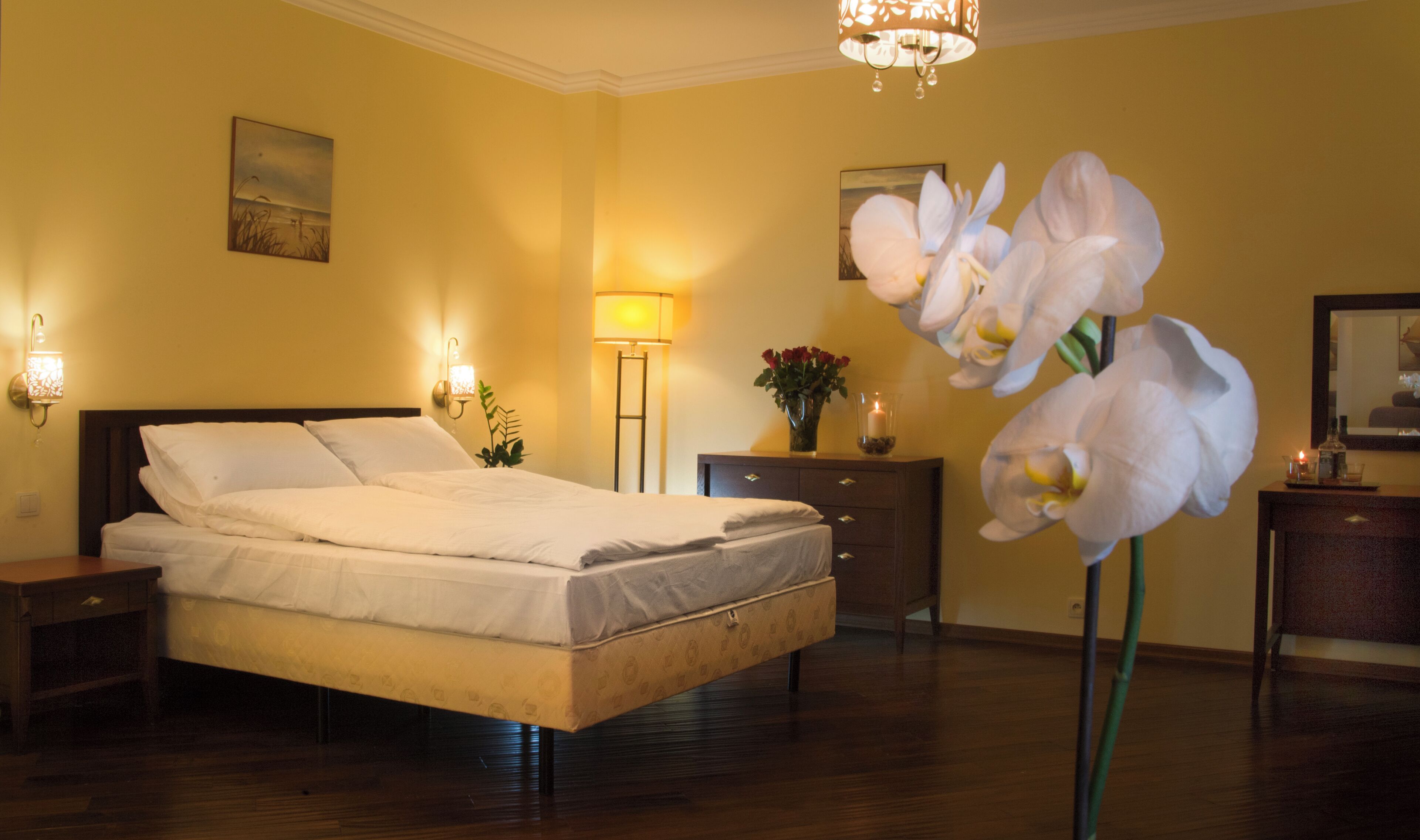 Habitación de hotel con cama, lámparas de noche, mesita de noche, cómoda, espejo y un florero con flores.