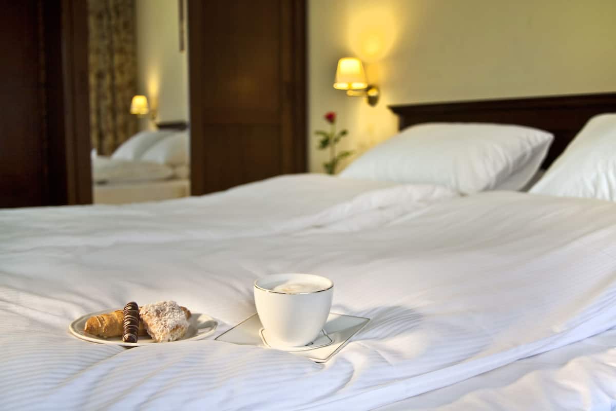 Un lit bien fait, une tasse de café et une pâtisserie sur une assiette, une lampe fixée au mur et un miroir qui reflète la pièce.