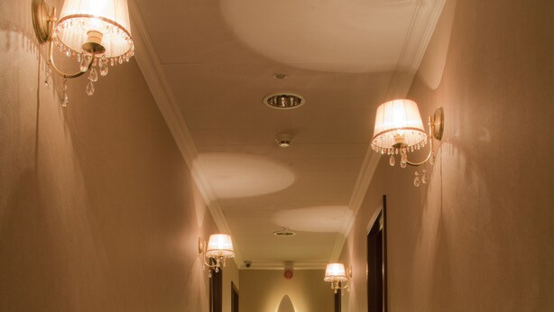 Een hotelgang met wandlampen, een gepolijste vloer en een centraal hanglamp.