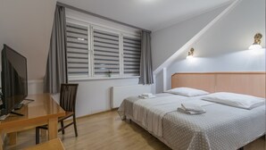 Quarto com cama, escrivaninha, cadeira, televisão e janela com cortinas.