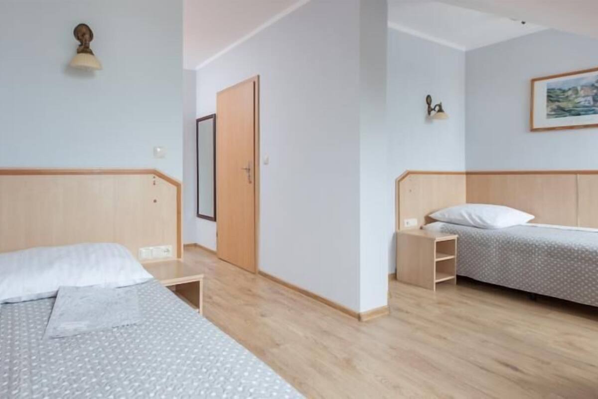 Une chambre avec deux lits, une tête de lit en bois, une table de chevet, une commode, une lampe fixée au mur et un tableau encadré.