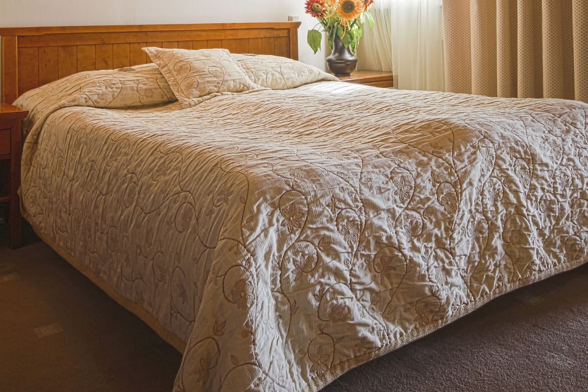 A bedroom with a wooden ceiling, a bed with a floral bedspread, two wall sconces, a vase with flowers, and a window with curtains.
