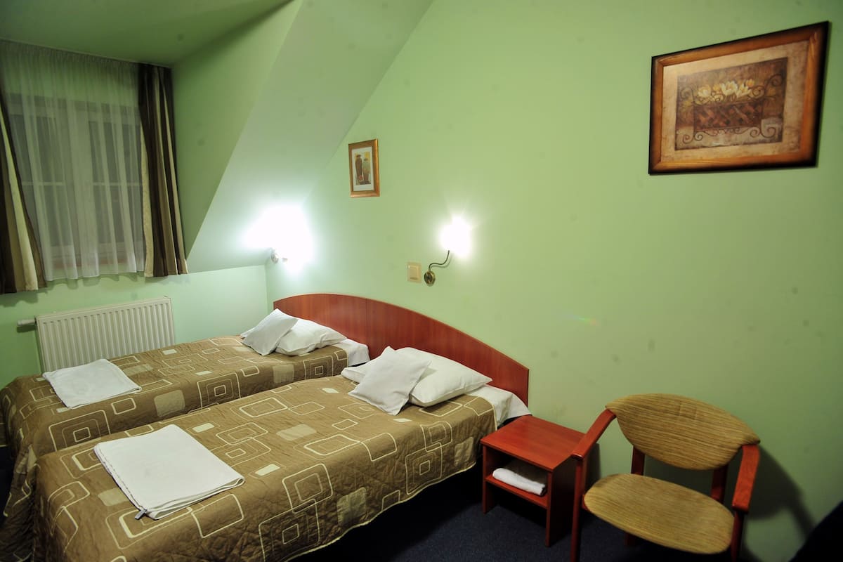 A hotel room with two beds, a chair, a small table, a radiator, a window with curtains, and a wall-mounted light fixture.