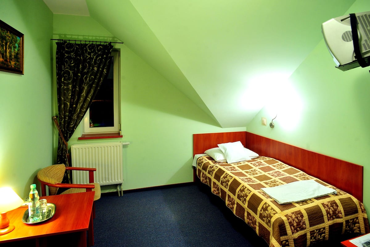 A small, single-bed room with a wooden headboard, a desk with a lamp and a bottle, a radiator, a window with curtains, and a ceiling-mounted projector.