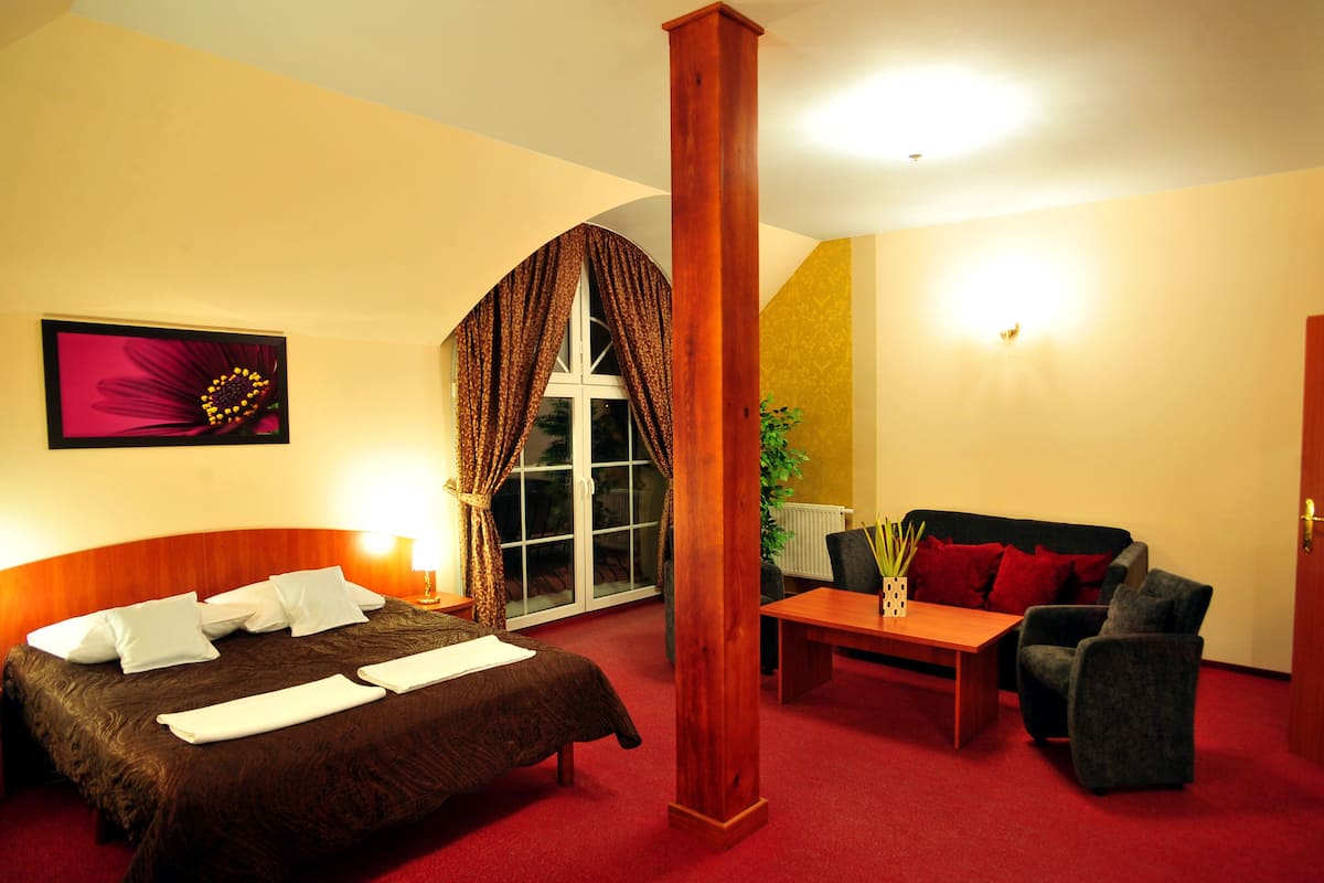 A hotel room with a bed, a sofa, a table, a window with curtains, a plant, and a painting on the wall.