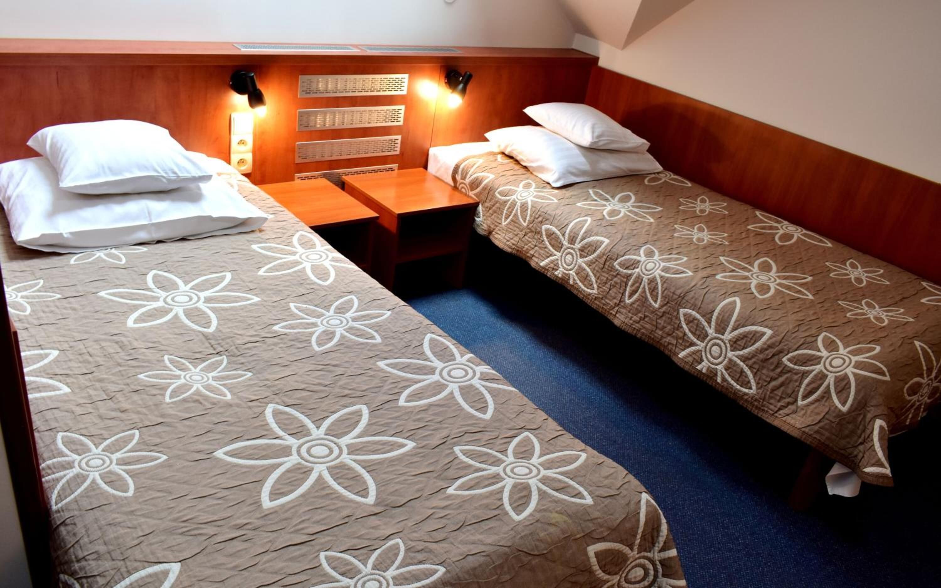 Habitación doble con dos camas individuales, cada una con una colcha de motivos florales. Tiene un cabecero de madera, una mesita de noche y un aplique de pared sobre las camas.