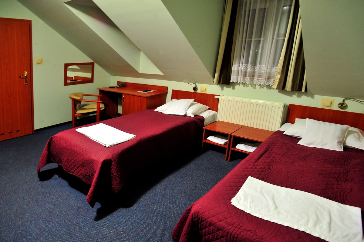 A hotel room with two beds, a desk, a chair, a window with curtains, and a radiator.
