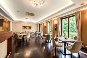 A spacious dining area with multiple tables and chairs, large windows with curtains, and a chandelier.