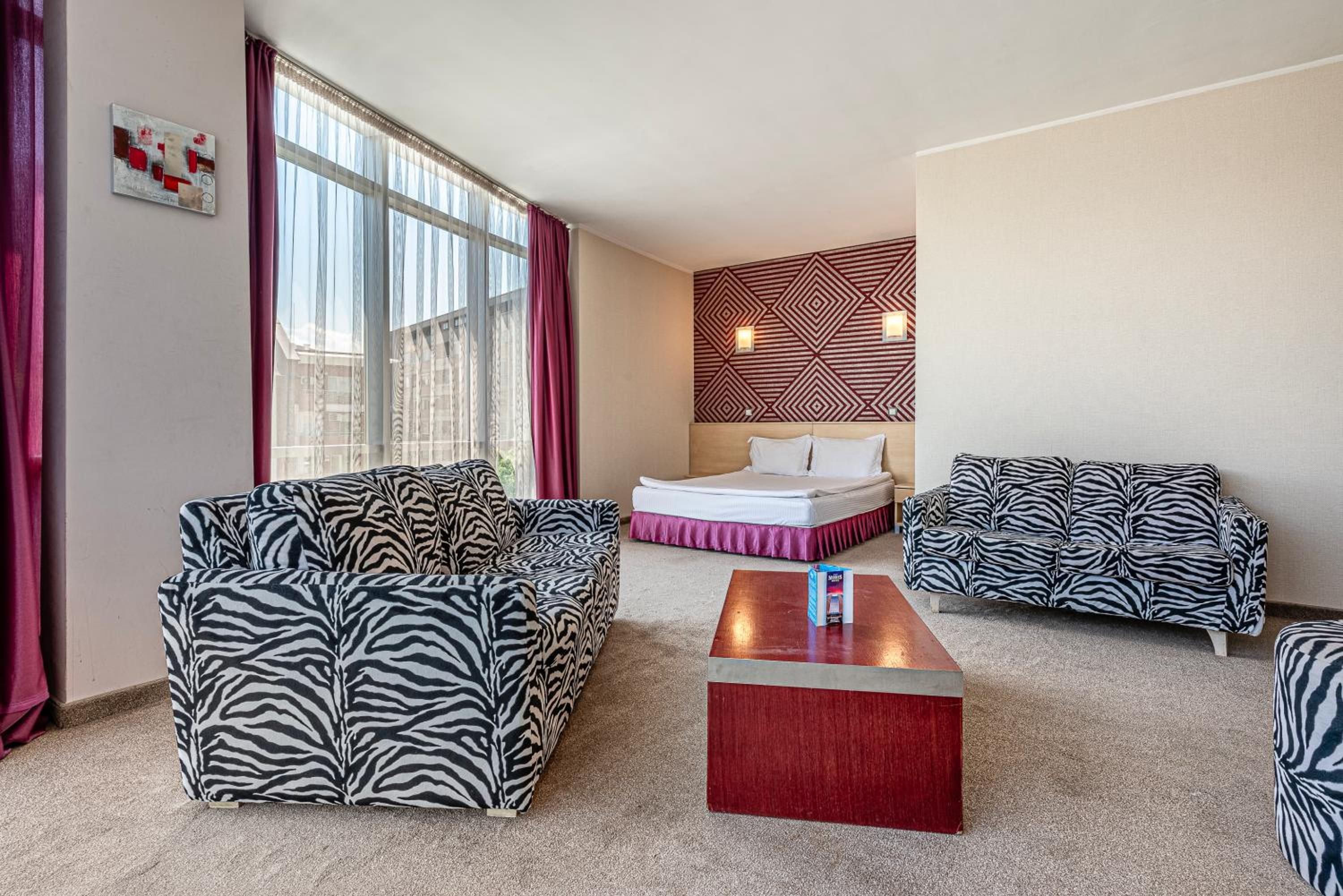 A hotel room with a zebra-patterned sofa set, a bed with a red accent, and a geometric-patterned wall.