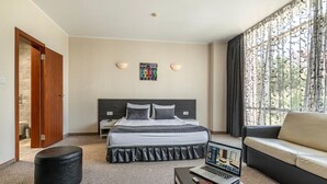 A hotel room with a bed, a sofa, a desk with a laptop, and a window with curtains.