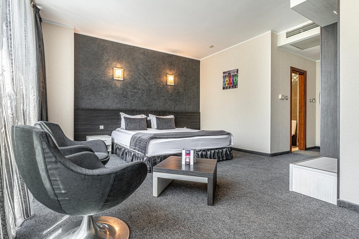 A hotel room with a bed, a chair, a small table, and a painting on the wall.