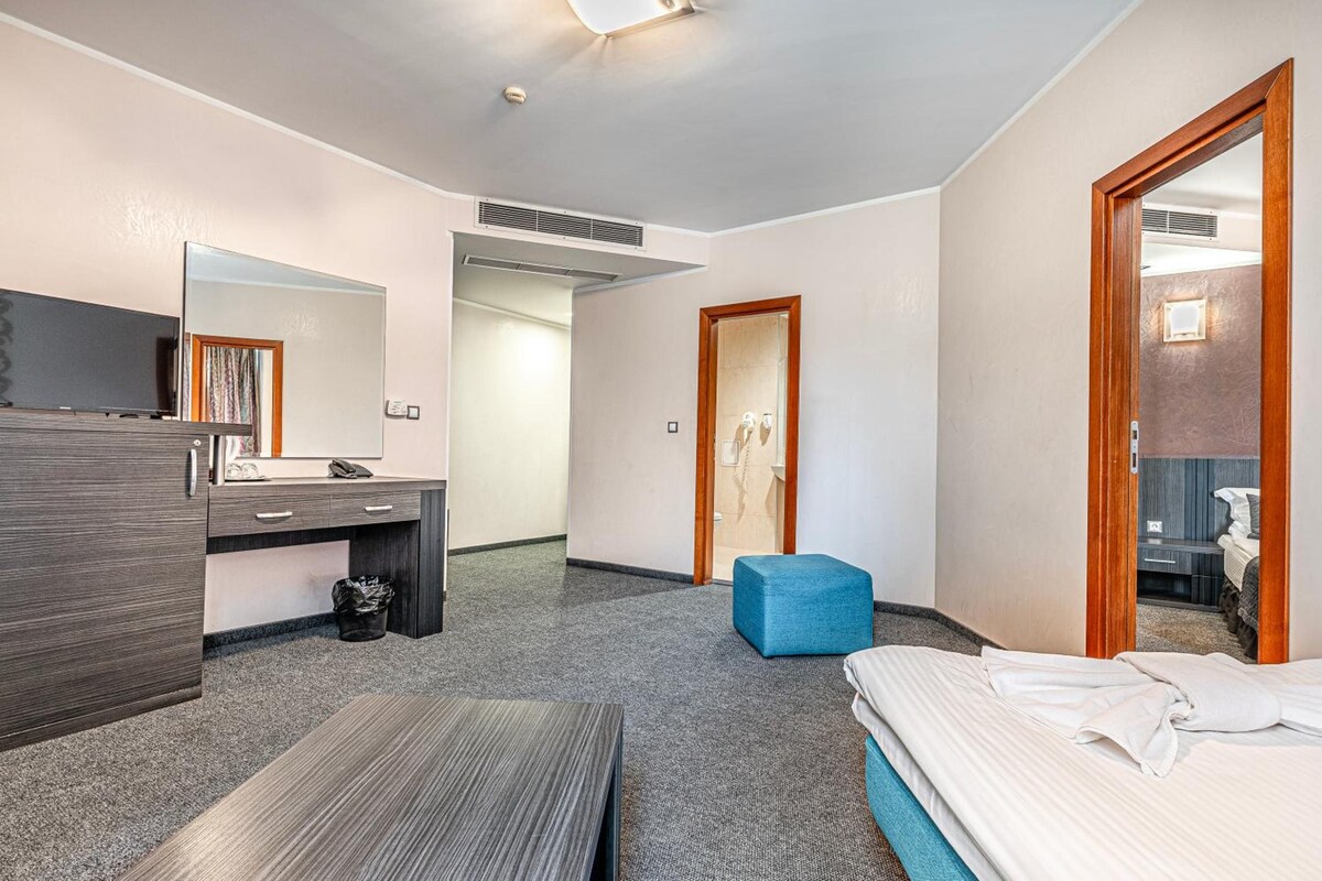 A hotel room with a bed, a desk with a mirror, a small blue ottoman, and a bathroom visible through an open door.